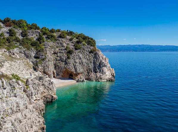 Una costa rocosa con un mar azul cristalino, una pequeña playa y un acantilado rocoso con vegetación escasa.