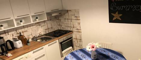 A compact kitchen with white cabinets, a wooden countertop, a sink, and a stove. There's a table with a blue tablecloth and a vase with flowers. A 'HELLO Summer' sign is on the wall.