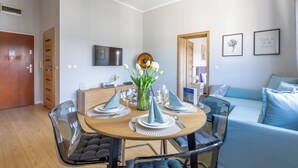 Uma sala de jantar moderna com uma mesa redonda posta para quatro pessoas, cadeiras de vidro transparente e um vaso com flores brancas. Um sofá azul e uma televisão são visíveis ao fundo.