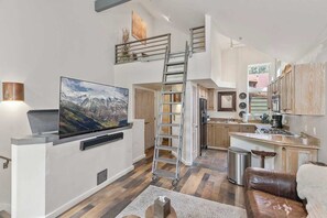 Una sala de estar moderna con televisor de pantalla plana, una mesa de centro de madera, un sofá de cuero marrón y una zona de cocina con armarios de madera y un lavamanos de acero inoxidable.