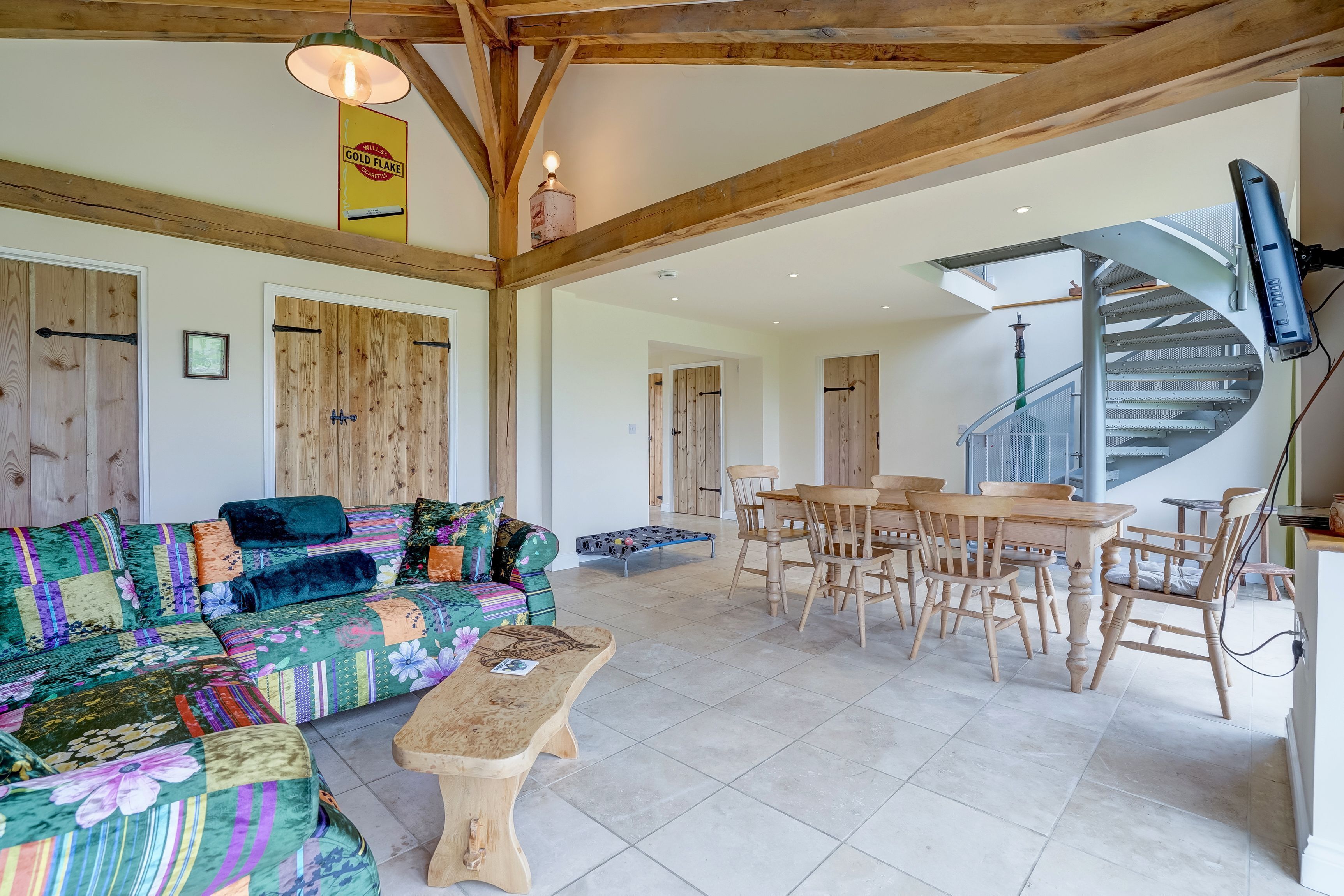 A spacious living area with a wooden ceiling, a colorful sofa, a wooden dining table with chairs, and a staircase leading to an upper floor.