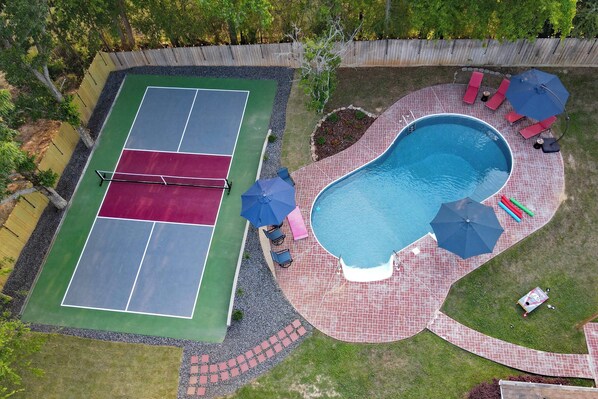Eine Vogelperspektive zeigt einen Garten mit Tennisplatz, Schwimmbecken und Sitzgelegenheiten im Freien.