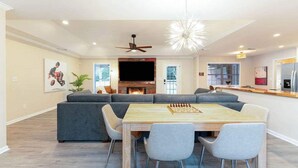 Un vaste salon comprenant un grand canapé d’angle gris, une table à manger en bois avec quatre chaises, une cheminée et un ventilateur de plafond.