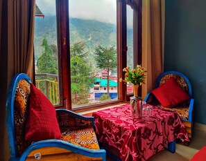 Habitación con vista a un paisaje montañoso, una mesa con un florero y dos sillas azules con cojines rojos.