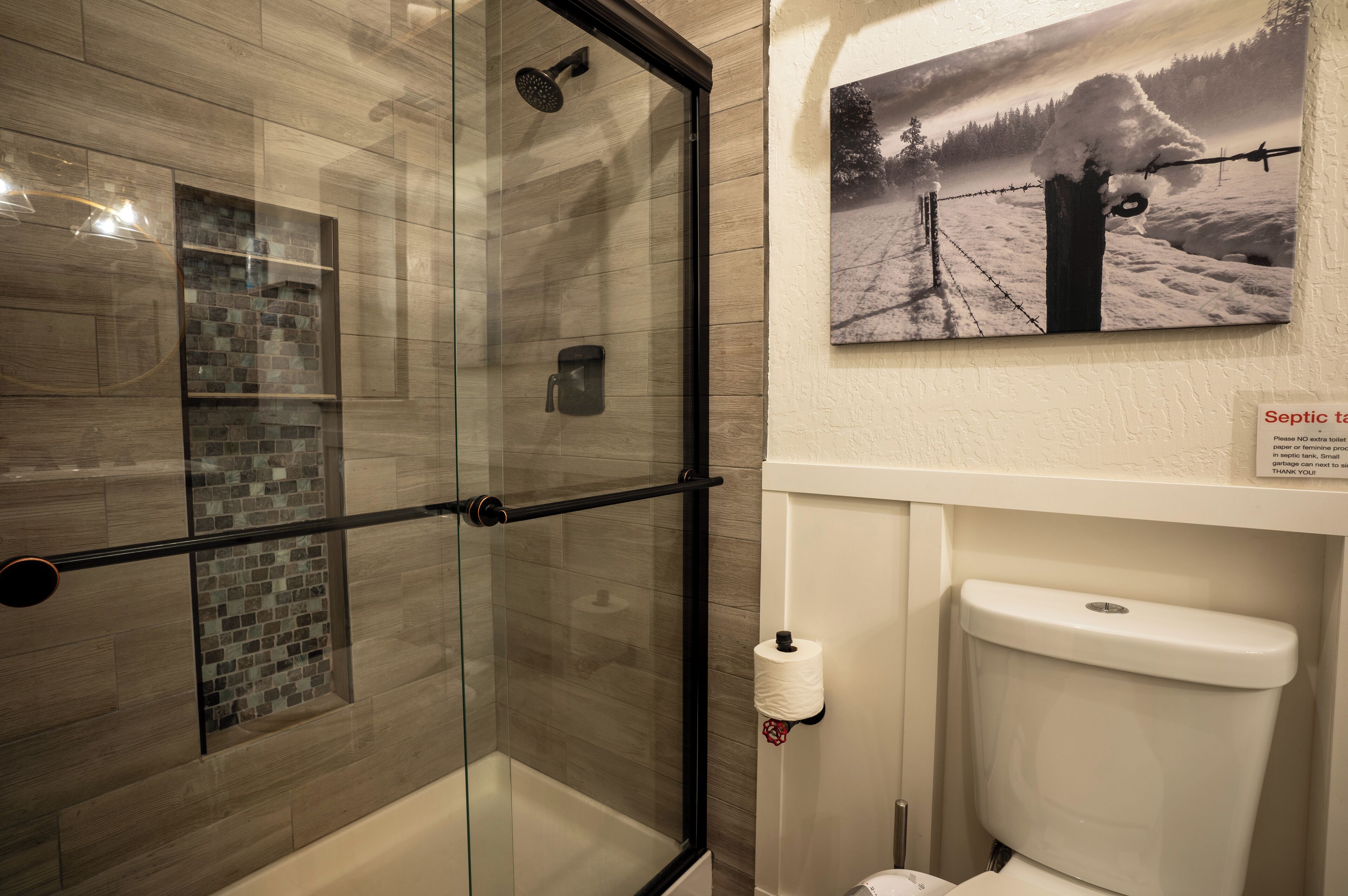 A bathroom with a glass shower enclosure, a mosaic tile wall, a toilet paper holder, and a framed picture of a snowy landscape.