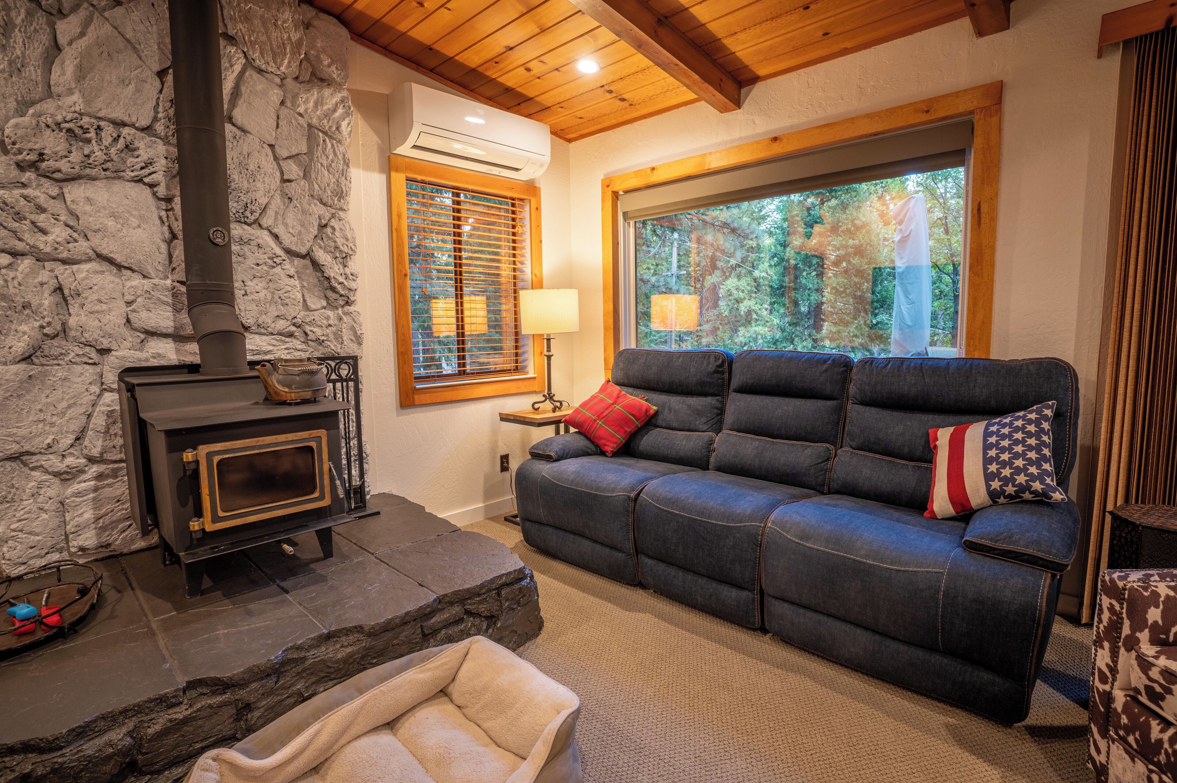 A cozy living room with a stone wall, a fireplace, a blue sofa with pillows, a window with blinds, and a lamp on a side table.