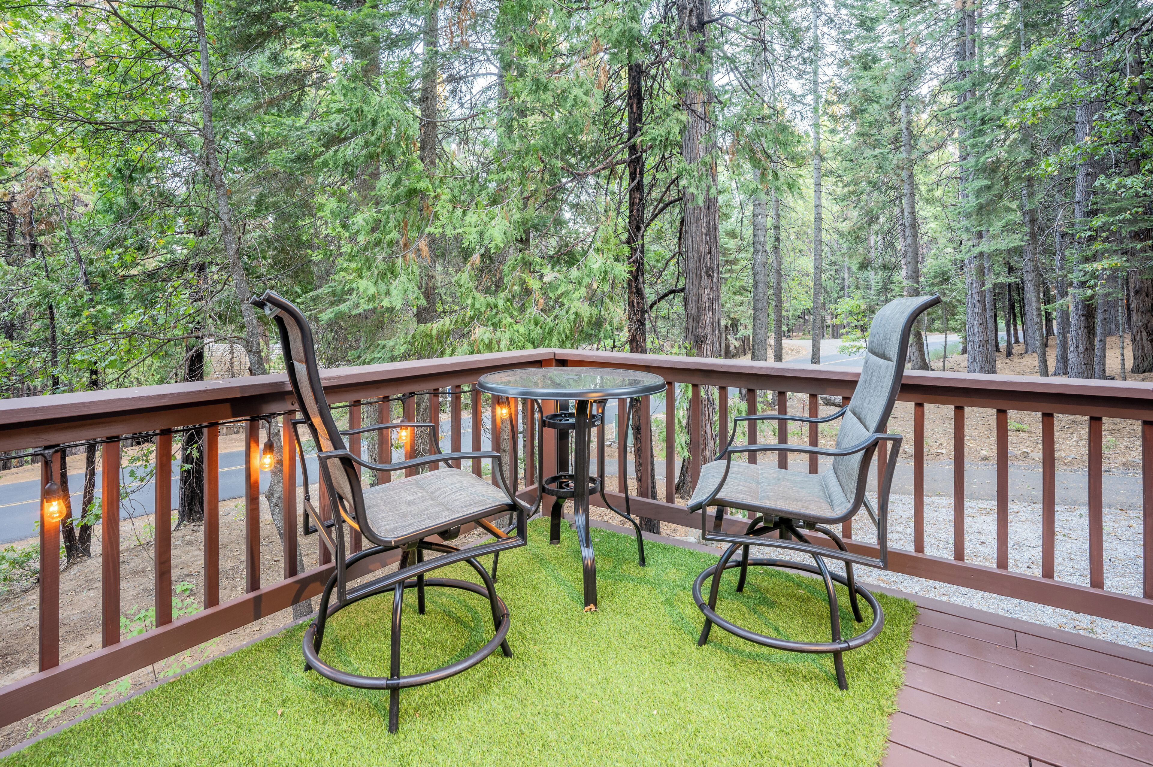 Eine Terrasse mit einem runden Tisch und zwei Stühlen, umgeben von einem Holgitter, vor einem Wald.