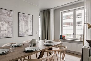 A dining area with a wooden table set for four, surrounded by chairs, with abstract art on the wall and a window with curtains. - Stylish Waterfront Apt Stockholm (Stockholms län)