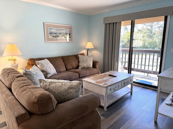 A living room with a brown sofa set, a white coffee table, and two lamps. There is a framed picture on the wall and a view of the outdoors through a sliding door.