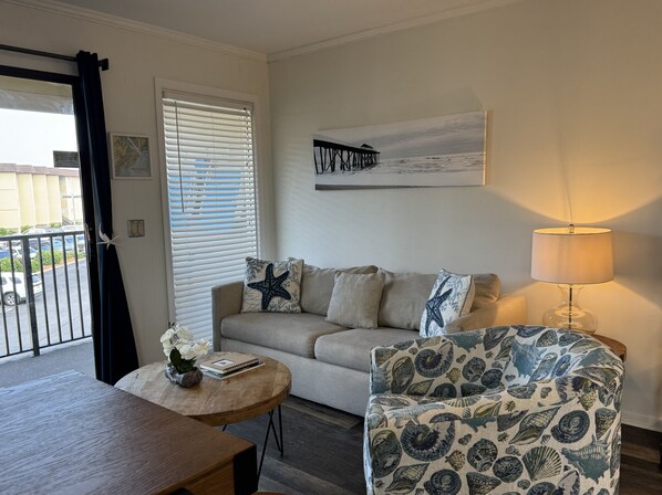 A living room with a sofa, a patterned armchair, a round wooden coffee table, a lamp, and a painting on the wall. - Ocean View - Ocean Villas B201-Ocean View - Professionally Managed by HHB&T Vacation Rentals<br>STR Permit# 35350 (Hilton Head Island)