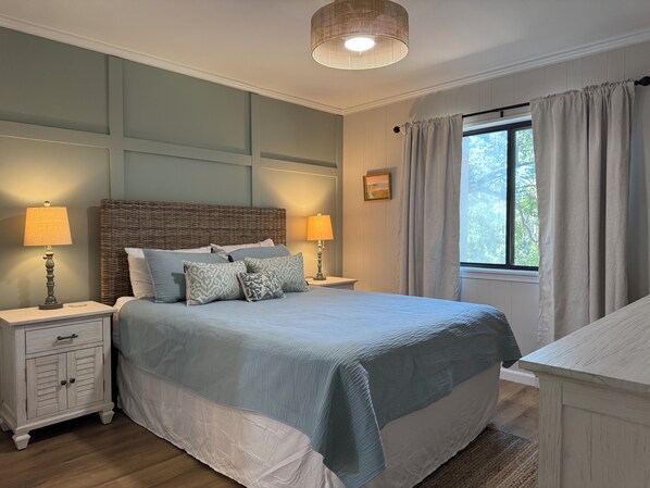 Une chambre à coucher avec un grand lit, deux tables de chevet, un luminaire suspendu, une fenêtre avec des rideaux et un tableau accroché au mur.