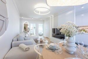 Una sala de estar moderna y luminosa, con un sofá, una mesa de comedor para dos personas y un televisor grande.