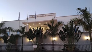 A building with a white facade, multiple flags, and a paved entrance area with palm trees. - Porto Golf Hotels (Kano)