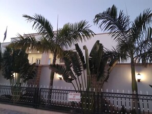 A building with a white facade, surrounded by a black metal fence, and palm trees in front. - Porto Golf Hotels (Kano)