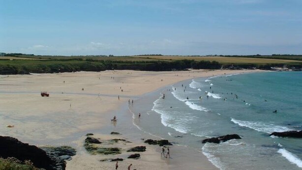 Une vaste plage où l’on voit des gens se promener et nager, entourée de verdure et sous un ciel dégagé.
