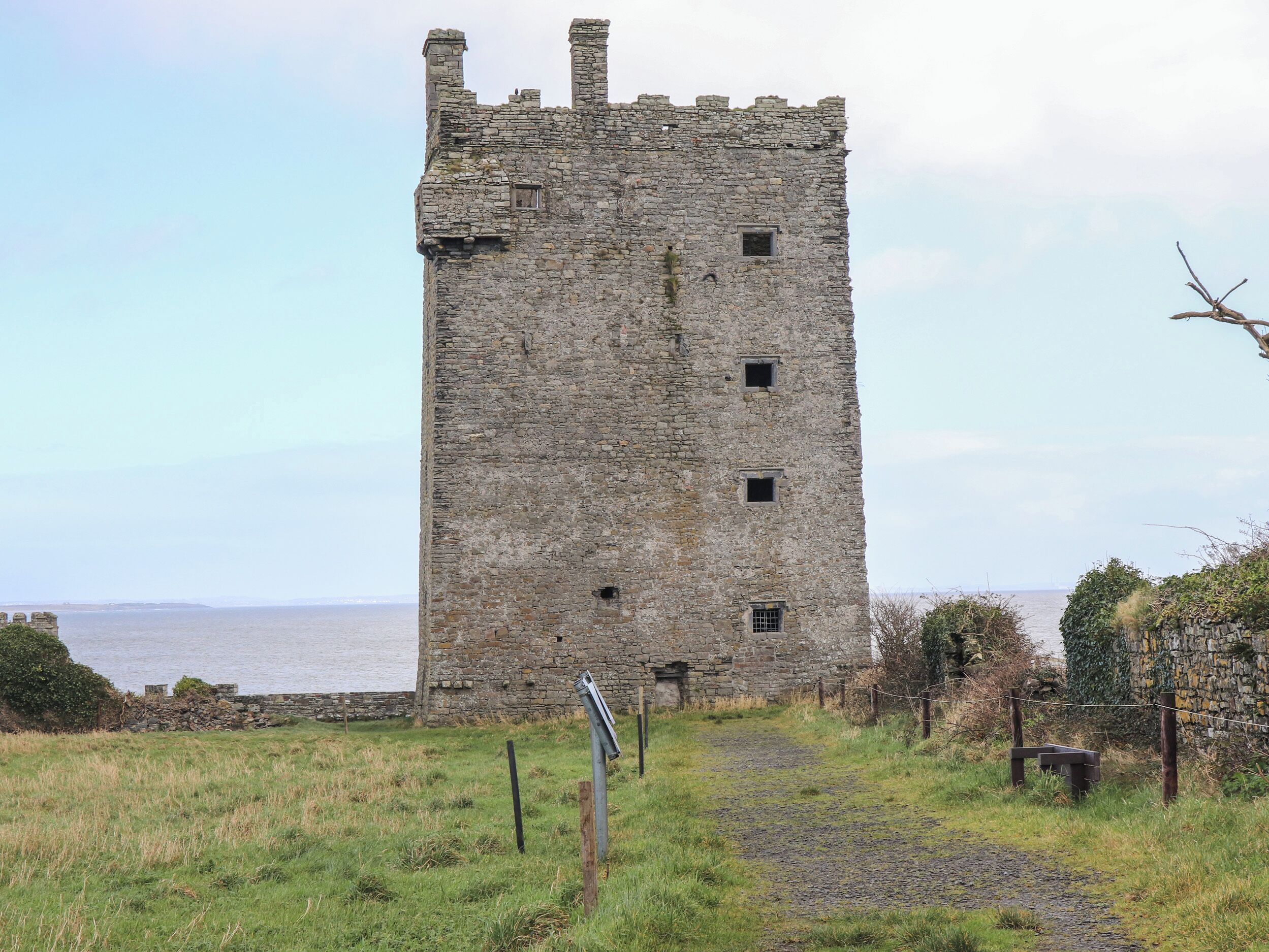 A historic stone tower with a narrow entrance, surrounded by grassy fields and a body of water in the background.