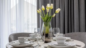 A dining table set with white plates, cups, and cutlery, a vase with white tulips, and a window with curtains in the background.