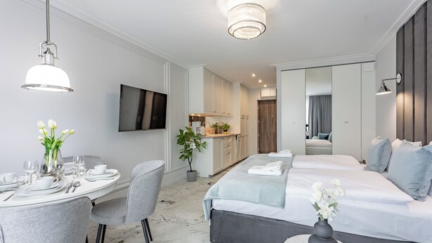 A modern hotel room with a dining area, a TV, a kitchen, and a large bed with a view of the city.