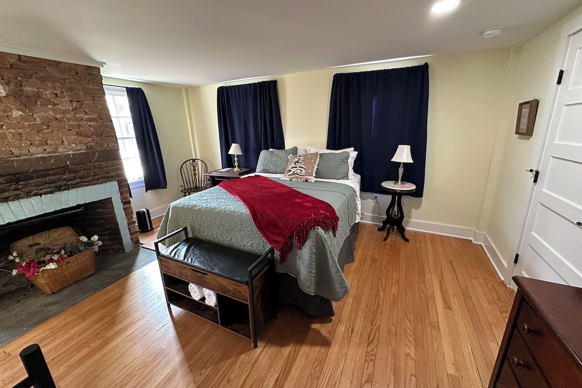 A bedroom with a brick fireplace, a bed with a red throw blanket, a wooden bench, and a small table with a lamp.