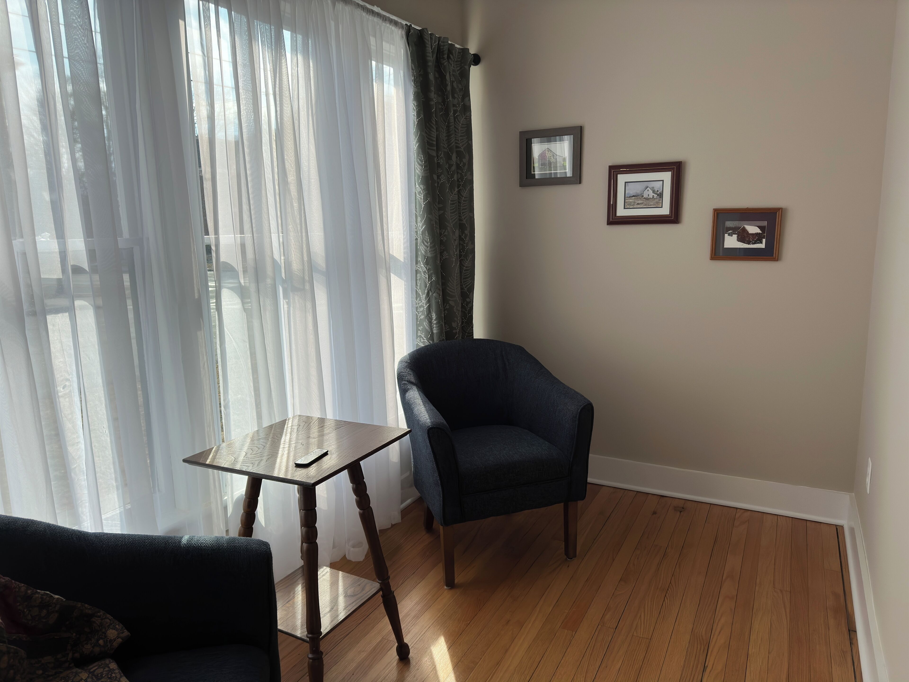 Una habitación con un sillón azul oscuro, una pequeña mesa de madera y un sofá. Hay tres cuadros enmarcados en la pared.