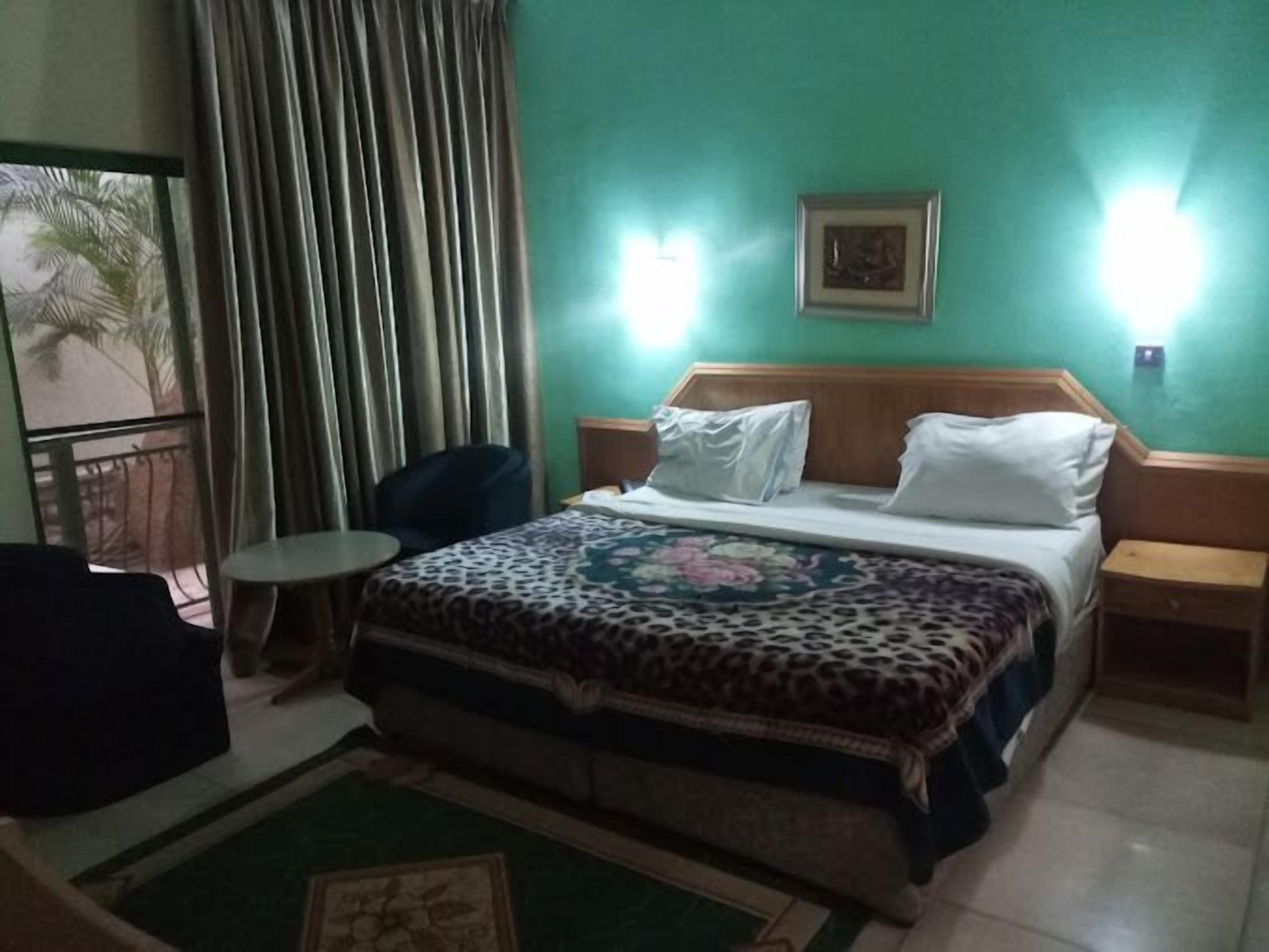 A hotel room with a bed, a chair, a small table, a window with curtains, and a wall-mounted light.