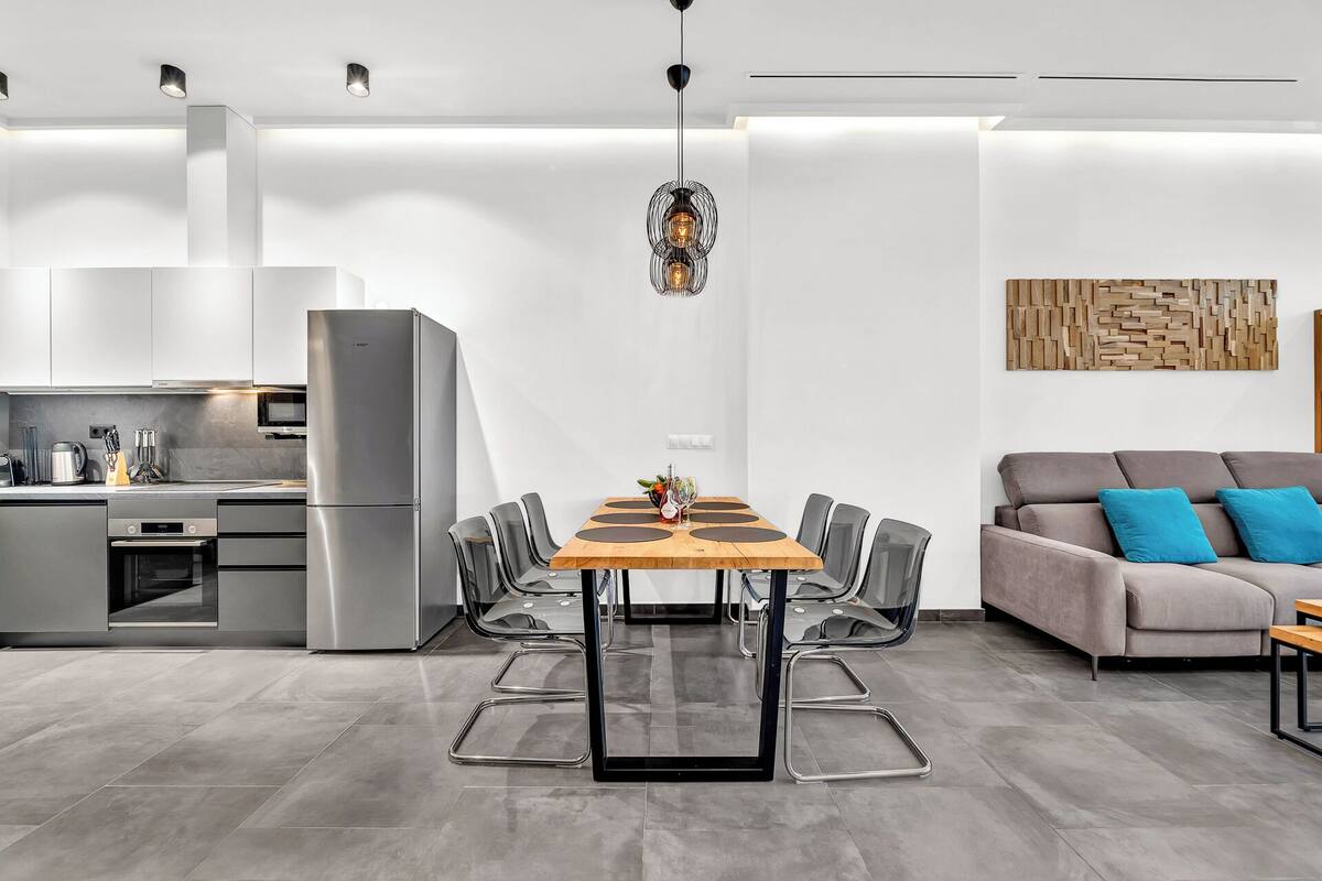 Una cocina moderna con refrigerador empotrado, una mesa de comedor con sillas y un sofá gris con cojines azules.