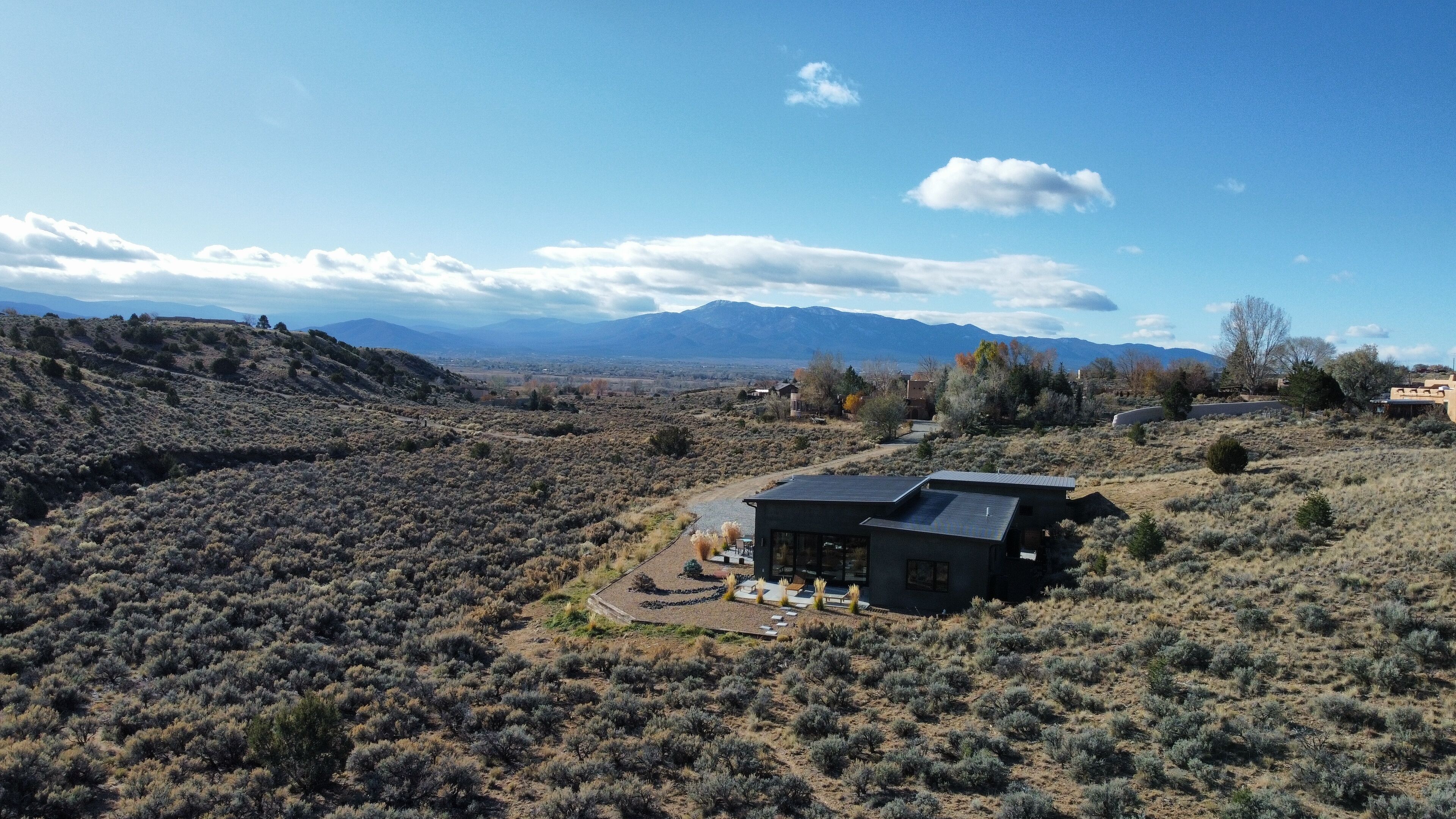 Une maison moderne dotée d’un toit recouvert de panneaux solaires, entourée d’un paysage aride et vallonné, où la végétation est rare et où l’on aperçoit des montagnes au loin.