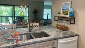 A modern kitchen with a marble countertop, a stainless steel sink, and a white refrigerator. There is a dining area with wooden chairs and a chandelier. A painting and a blue vase are on the wall above the kitchen counter. - Bocilla Island Club Unit C-13 (Bokeelia)