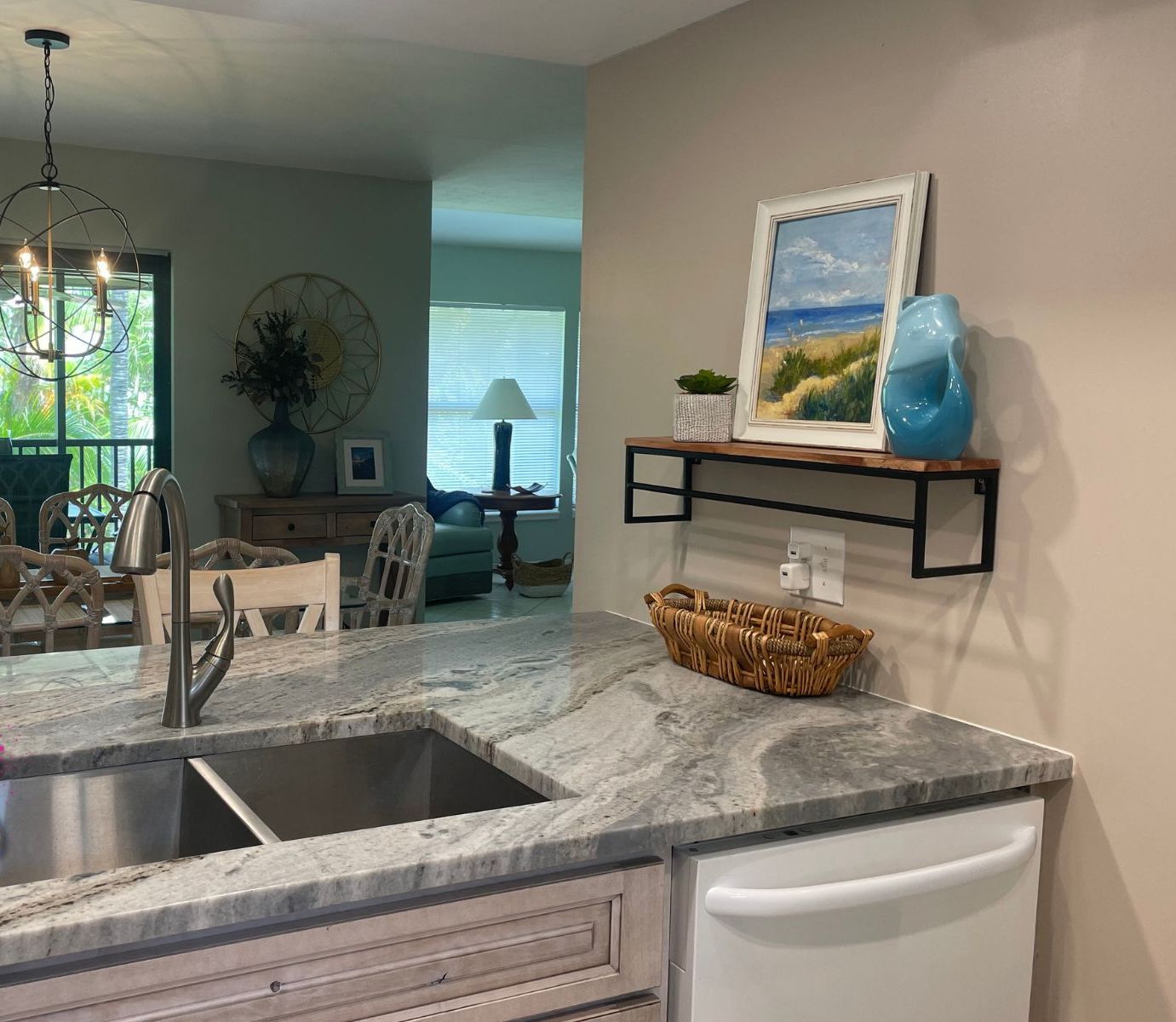 A modern kitchen with a marble countertop, a stainless steel sink, and a white refrigerator. There is a dining area with wooden chairs and a chandelier. A painting and a blue vase are on the wall above the kitchen counter.