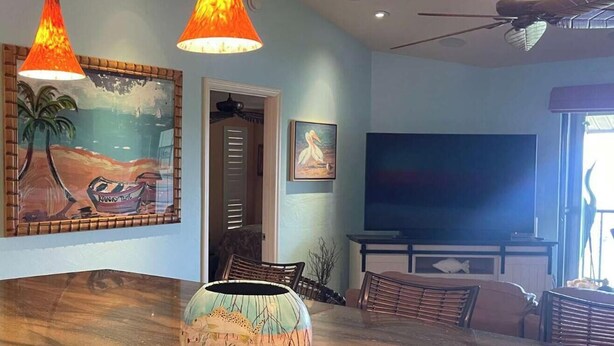 A spacious living room with a large wooden dining table, two hanging orange lamps, a painting of a tropical scene, and a television in the background. - Captains Harbor Island Hide Away (Bokeelia)