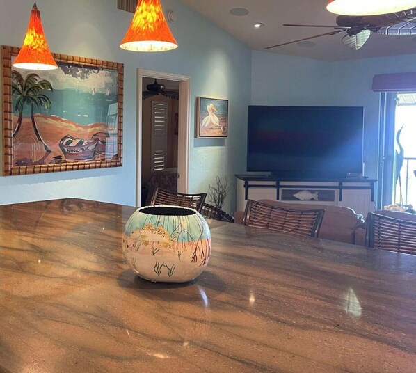 A dining area with a large table, two hanging lamps, a painting, and a television.