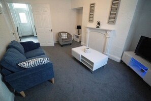 A living room with a blue sofa, a white coffee table, a grey armchair, a small side table with a lamp, a fireplace, and a TV on a stand.