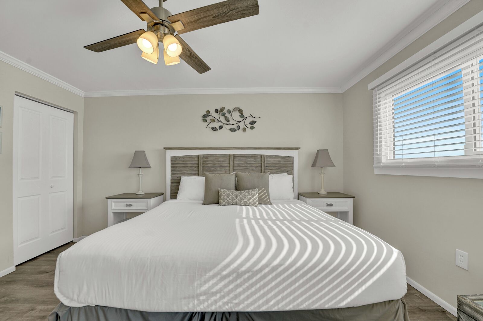 A bedroom with a large bed, two bedside tables with lamps, a ceiling fan, a decorative wall piece, and a window with blinds.