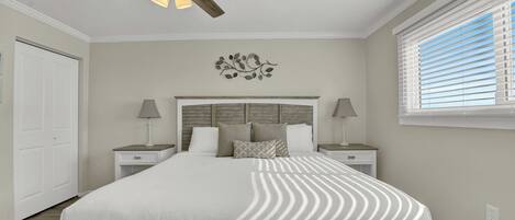 A bedroom with a large bed, two bedside tables with lamps, a ceiling fan, a decorative wall piece, and a window with blinds.