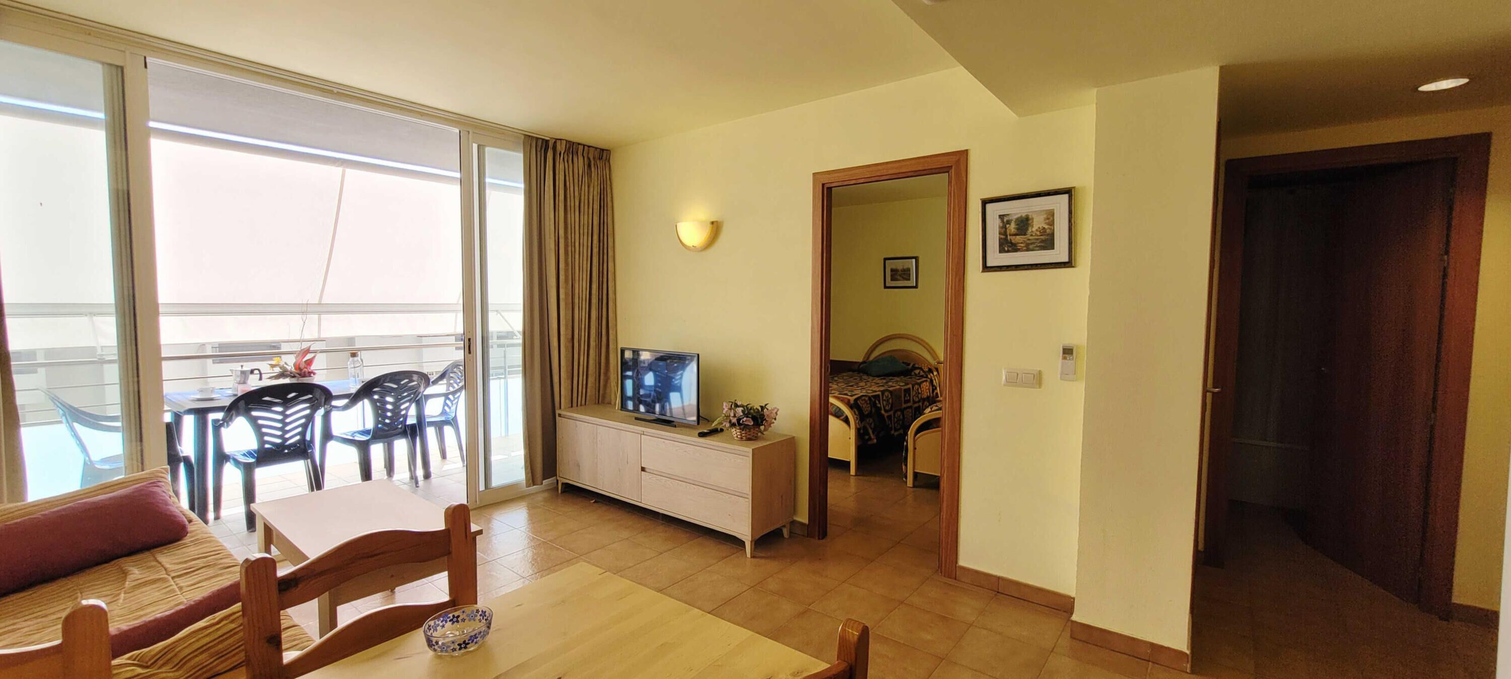 Une chambre d’hôtel avec un balcon, un coin repas avec une table et des chaises, un téléviseur posé sur un meuble, et un lit recouvert d’une couvre-lit à motifs.