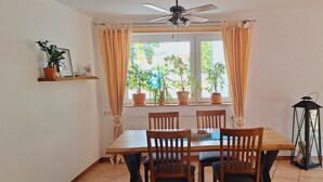Un coin repas avec une table en bois, quatre chaises, un ventilateur de plafond, des plantes en pot sur le rebord d’une fenêtre et un téléviseur fixé au mur.
