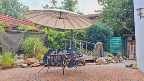 Une terrasse avec une table et des chaises, un grand parasol, et un jardin agrémenté de plantes et de rochers.
