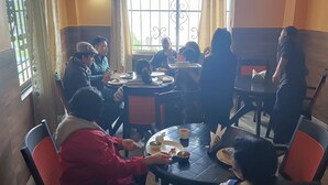 Un groupe de personnes est réuni autour d’une table ronde, et elles déjeunent ou dînent en admirant le paysage à travers une fenêtre ornée de barreaux en métal.