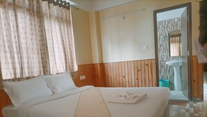 Un lit bien fait, avec du linge de lit blanc et un plaid beige, une tête de lit en bois, et une vue sur une salle de bain avec un lavabo et un miroir.