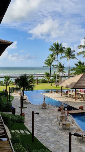 A resort pool area with a view of the ocean, palm trees, and outdoor seating. - La Fleur Polinésia Residence - Muro Alto (Ipojuca)