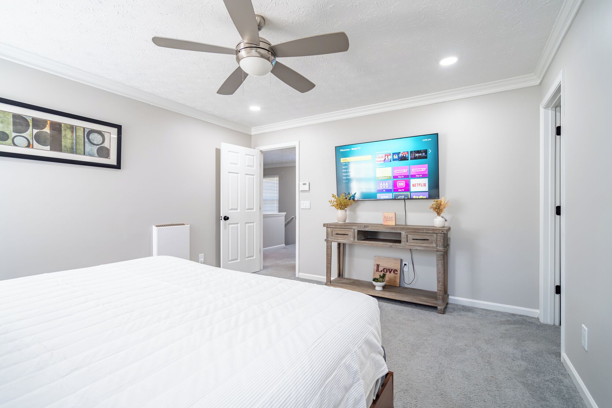 A bedroom with a bed, a ceiling fan, a TV mounted on the wall, and a small console table with decorative items.