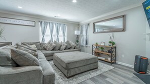A modern living room with a sectional sofa, a small ottoman, and a wooden coffee table. There is a large mirror on the wall, a floor lamp, and a plant on a wooden shelf. - Charming 3-bedroom house with AC, WiFi in terrific Lithia Springs (Lithia Springs)