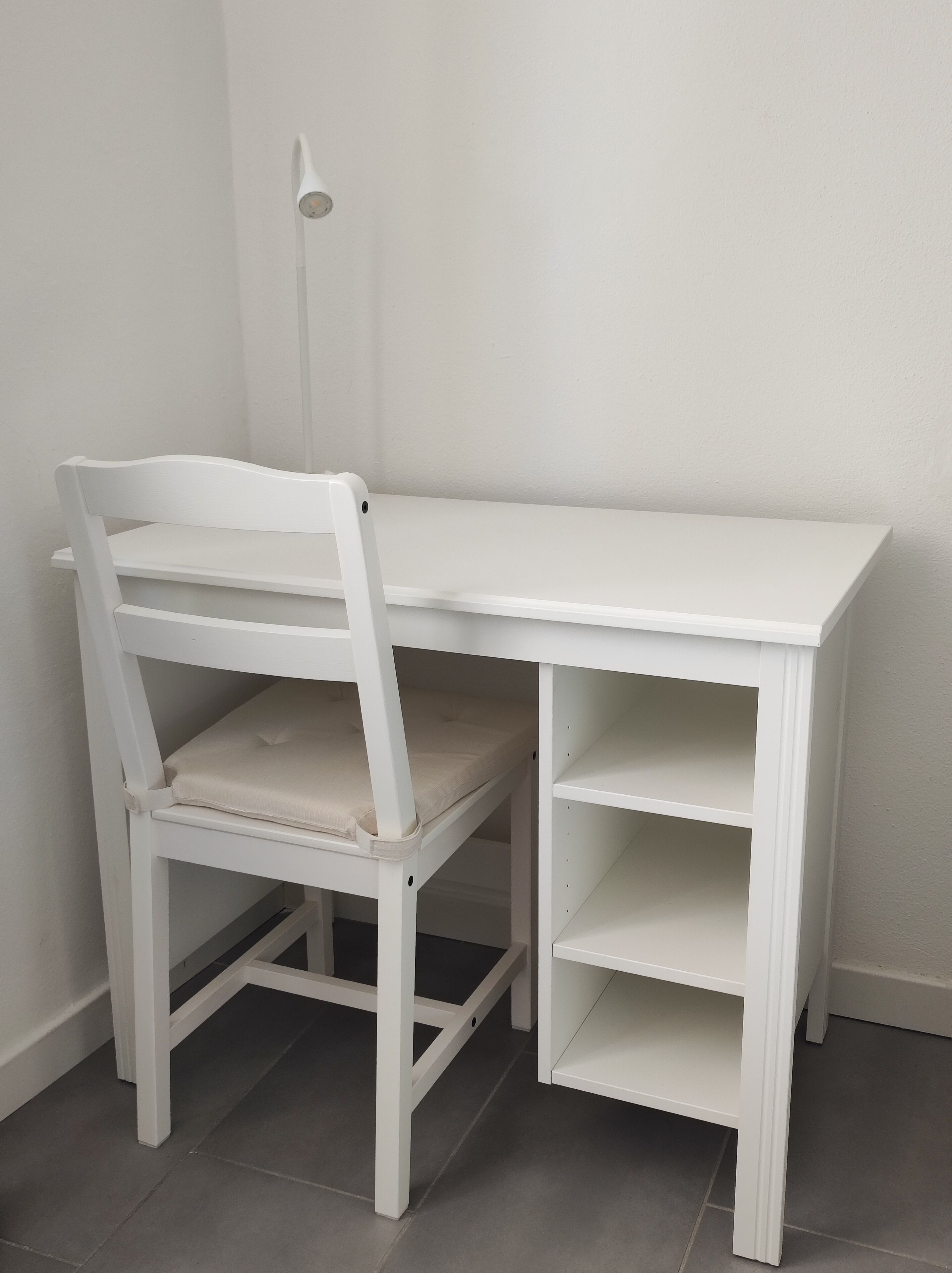 Un escritorio blanco con una silla, una estantería y una lámpara.
