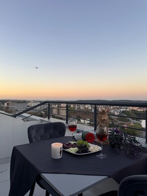 A table set with a plate of grapes, a glass of red wine, and a candle on a balcony overlooking a cityscape at sunset. - BZN AİRPORT HOTEL (Arnavutköy)
