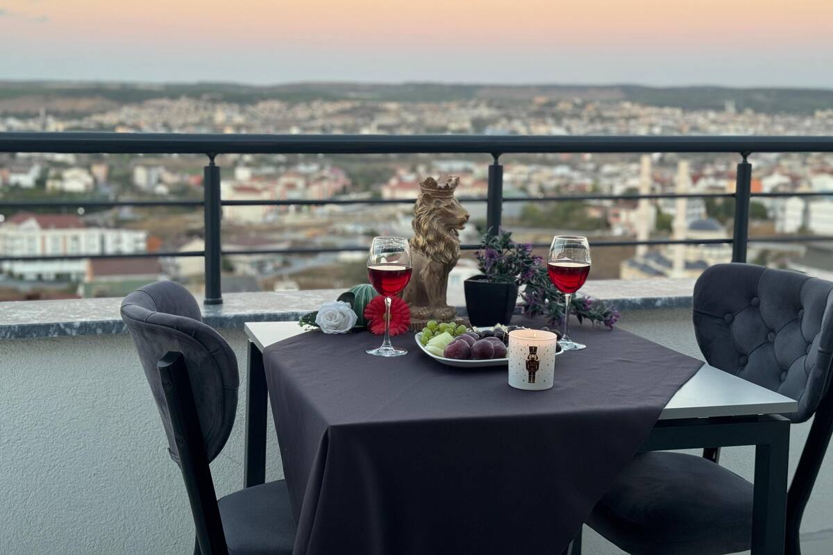 A table set with a wine glass, a candle, and a plate of grapes on a balcony overlooking a cityscape.
