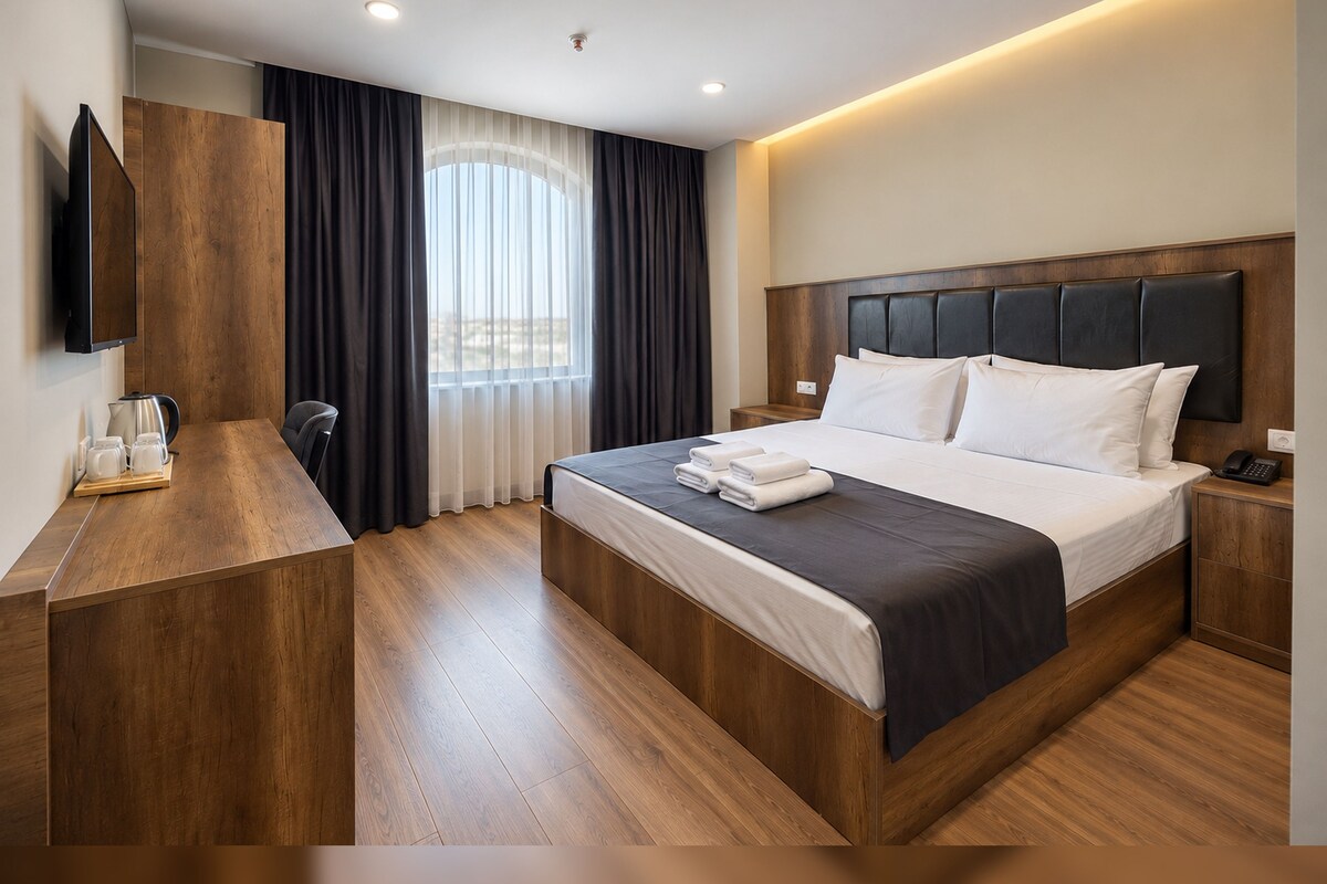A modern hotel room with a large bed, a bedside table, a desk with a coffee maker, and a TV mounted on the wall.