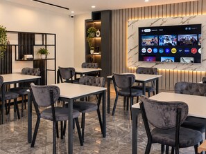 A modern dining area with multiple tables and chairs, a wall-mounted TV displaying various channels, and a decorative shelf with plants. - BZN AİRPORT HOTEL (Arnavutköy)