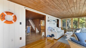 A spacious living area with a wooden ceiling, a white wall with a life preserver, a staircase, and a dining area with natural light. - Point Comfort On Lake Winnisquam (Sanbornton)