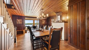 A spacious dining area with a long wooden table and chairs, a staircase with wooden railings, and a view of the outdoors through large windows. - Point Comfort On Lake Winnisquam (Sanbornton)