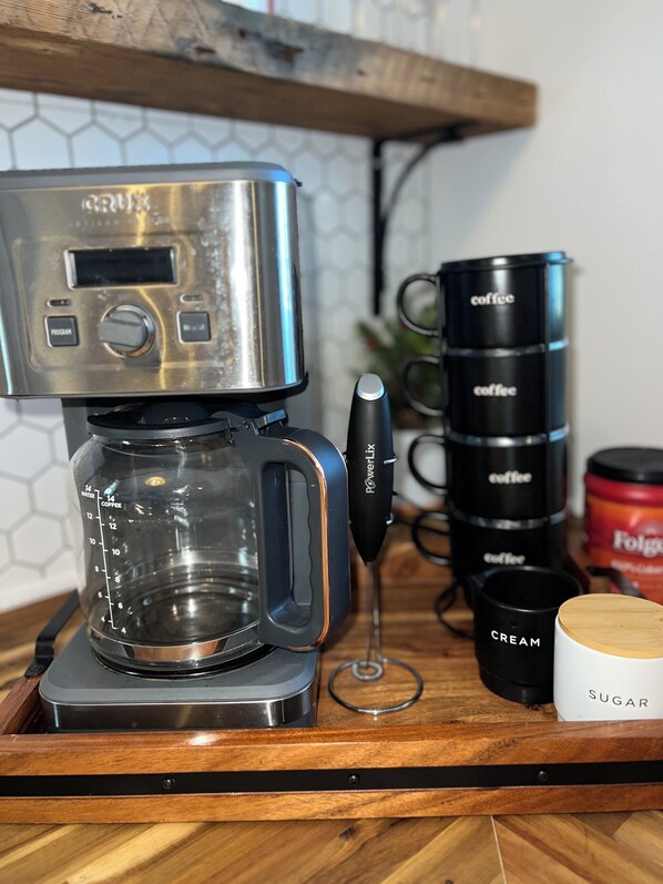 Una cafetera sobre una encimera de madera, con jarra de vidrio, un molinillo de café y tazas de café etiquetadas.
